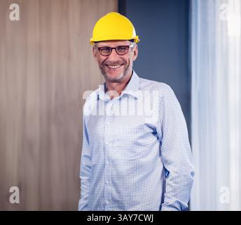 Senior man standing in office with wood wall and curtain, wearing yellow hard hat and eyeglasses Stock Photo