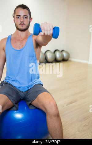 young adult man sitting on stability ball in gym studio, lifting blue dumbbell with silver balls Stock Photo