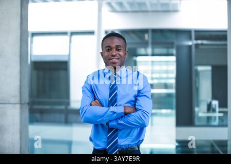 Young African American man standing arms crossed in office lobby, wearing blue shirt striped tie Stock Photo