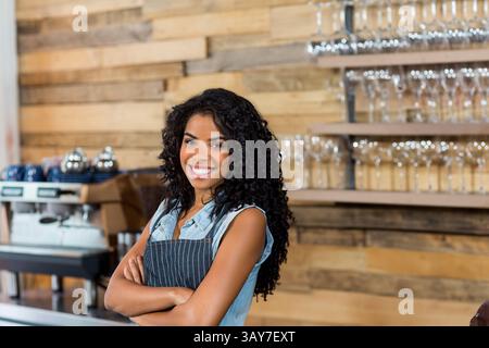 Young adult woman standing behind wooden bar counter in coffee shop, smiling near espresso machine Stock Photo