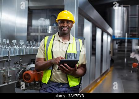 African American man overseeing glass bottles conveyor in factory using tablet and wearing hard hat Stock Photo