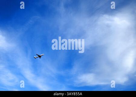 Airplane flying in a clear blue sky with wispy white clouds. The aircraft is positioned in the left section of the image, giving a sense of motion and Stock Photo