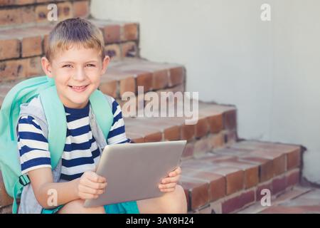 Boy sitting on outdoor brick stairs, holding silver tablet with mint green backpack, copy space Stock Photo
