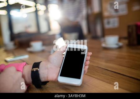 Person's left hand holding white smartphone over wooden table in café, showing coffee cups Stock Photo