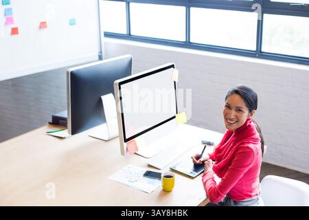 Young Chinese woman creating art on drawing tablet in office, with dual monitors and sticky notes Stock Photo