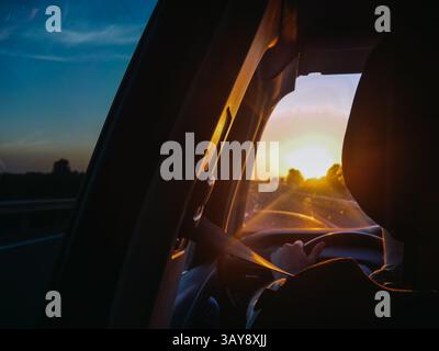 A view from the interior of a car, focusing on the drivers hands on the steering wheel and the warm glow of the setting sun visible through the windsh Stock Photo
