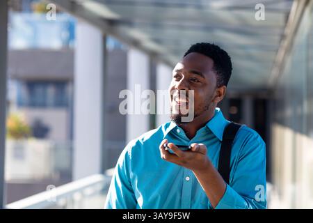 Young African American man using smartphone with backpack on covered outdoor walkway, smiling Stock Photo