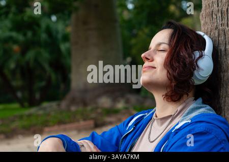 Relaxed woman enjoying music with closed eyes and wireless headphones while sitting against a tree in a peaceful park setting, embracing nature's tran Stock Photo