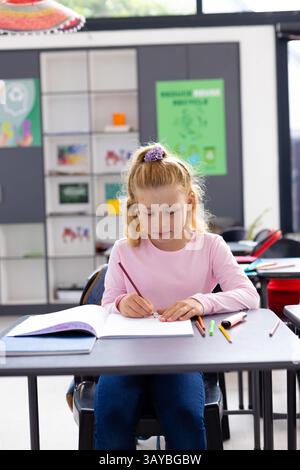 Girl child drawing in notebook at school desk in bright classroom, with colored pencils scattered Stock Photo