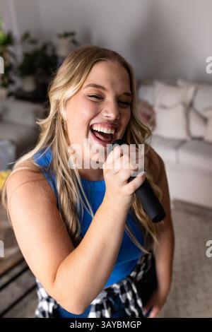 Young adult woman singing while standing in living room, holding handheld microphone and smiling Stock Photo