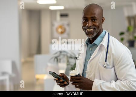 African American male doctor smiling, holding smartphone and coffee cup in clinic, copy space Stock Photo