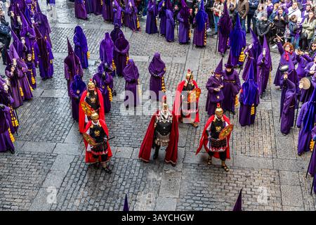 Brotherhood members in Roman costume stand in formation among purple-robed nazarenos on the Calle del Fuero in Cuenca, Castilla-La Mancha, Spain. Red-plumed helmets, golden breastplates and red capes mark their uniforms. They accompany the Good Friday procession Camino del Calvario, known as Las Turbas. During Semana Santa in Cuenca, 33 Hermandades take part in eleven processions through the city during Holy Week. Stock Photo