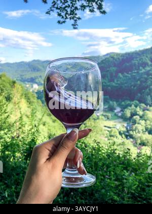 Hand holding a glass of red wine against a stunning outdoor backdrop of lush green hills and a beautiful sky. Stock Photo