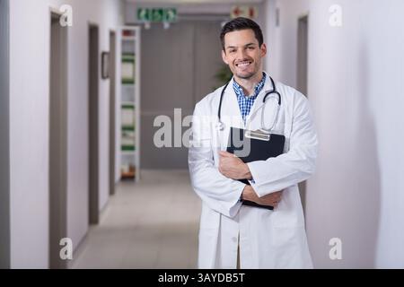 Asian male doctor standing in hospital corridor holding clipboard with stethoscope, copy space Stock Photo
