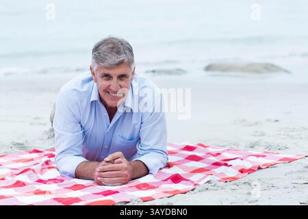 Senior man lying on red and white checkered picnic blanket on sand beach, smiling at camera Stock Photo