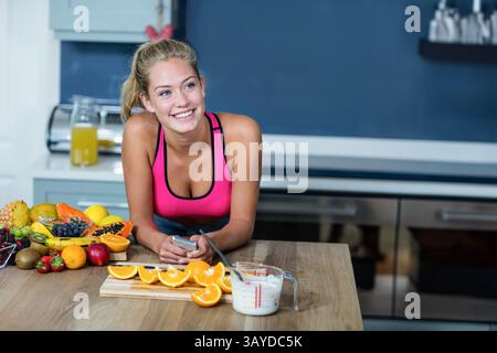 Woman leaning on countertop in modern kitchen, holding smartphone near cutting board with fruits Stock Photo