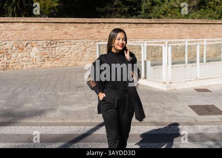A fashionable woman in a chic black outfit confidently walks and talks on her phone in a vibrant urban setting Stock Photo