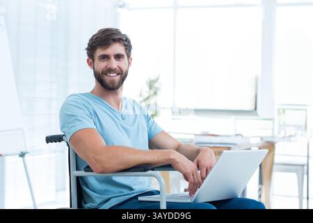 Young adult man working on laptop at modern office space, displaying wheelchair and whiteboard Stock Photo