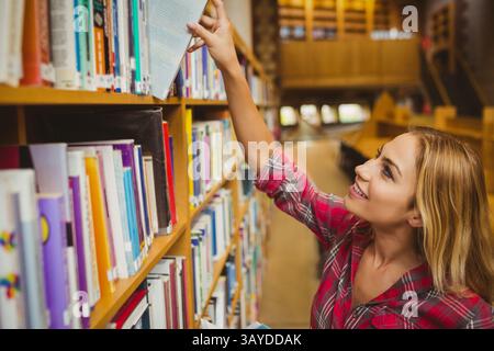Female student reaching for hardcover book on wooden shelf in library aisle by tables, copy space Stock Photo