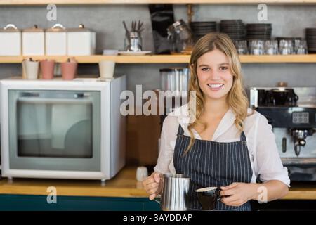 Female barista smiling behind counter in coffee shop holding milk frothing pitcher and coffee cup Stock Photo