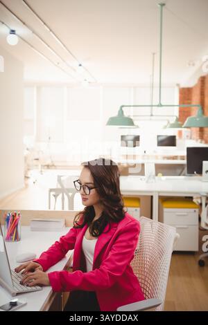 Asian young woman typing at laptop in modern sunlit open plan office, with colored pencil holder Stock Photo