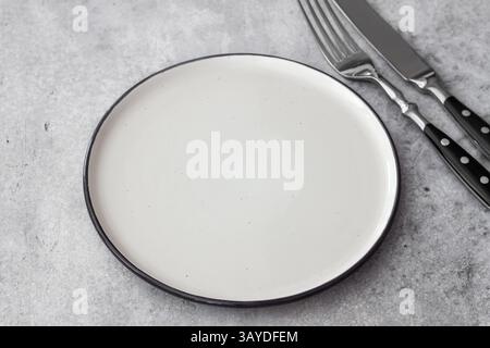 Minimalist top view of an empty white ceramic plate with knife and fork on gray stone background Stock Photo