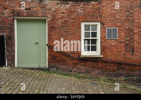 Old red brick wall on steep cobbled street with single green exterior door, single paneled window, single air vent and metal bannister or handrail Stock Photo