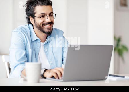 Smiling Indian Writer Using Laptop Computer, Sitting At Desk In Home Office Stock Photo