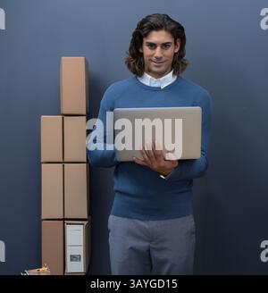 Middle Eastern young adult man holding silver laptop standing in office space with stacked boxes Stock Photo