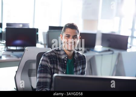 Young man working on desktop computer in modern office, smiling at camera with computer monitor Stock Photo