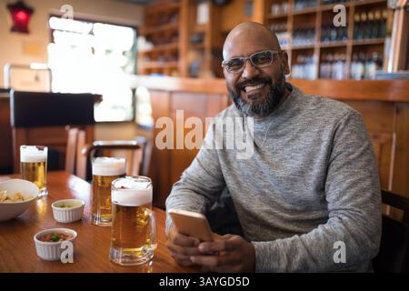 Indian man enjoying beer and chips with salsa at wooden pub table, smiling and using smartphone Stock Photo
