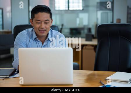 Asian man sitting at wooden desk in open-plan office, working on silver laptop with white notepad Stock Photo