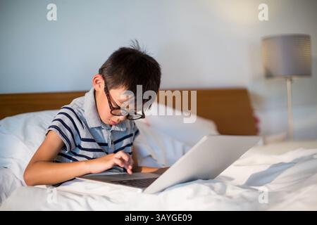 Asian boy child sitting on bed using silver laptop in bedroom with eyeglasses and lamp Stock Photo