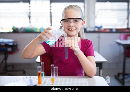 Schoolgirl pouring blue liquid from beaker into test tube at classroom desk with safety goggles Stock Photo