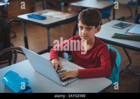 Boy typing on silver laptop at school desk in classroom, with blue backpack and pencil case Stock Photo