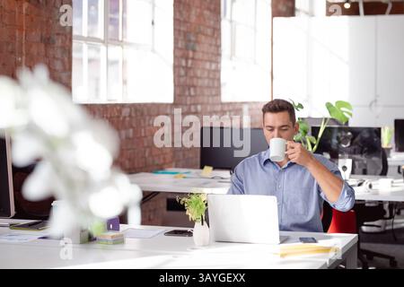 Young adult man drinking coffee and working on laptop at modern open-plan office desk, copy space Stock Photo