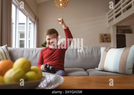 Boy playing video game with black controller while sitting on gray sofa in modern living room Stock Photo