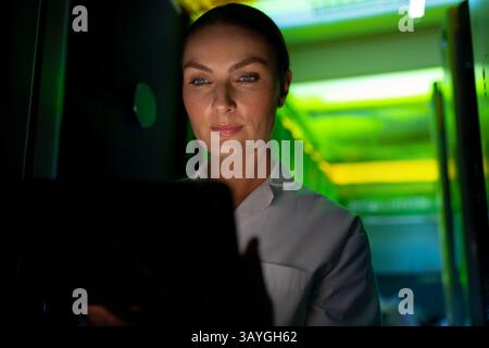 Woman checking tablet in data center corridor, surrounded by server racks and LED lights Stock Photo