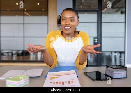 African American woman gesturing with hands at desk in modern office, using tablet and chart Stock Photo