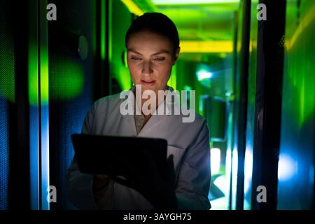 Woman using tablet monitoring LED lights and cables in server aisle at data center, copy space Stock Photo