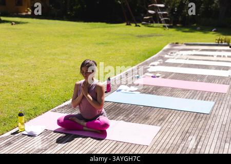 young pregnant woman practicing yoga on pink mat in outdoor wooden deck garden, with water bottles Stock Photo