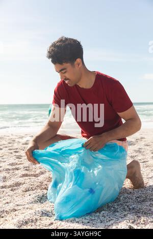 Man kneeling on sandy beach at shoreline, collecting litter with large light blue plastic bag Stock Photo