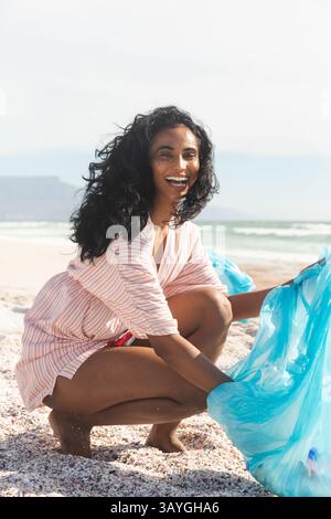 Mid-adult bi-racial woman crouching on beach by water, picking up bright blue plastic bag Stock Photo
