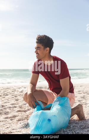 Man kneeling on sandy beach near ocean waves, holding blue plastic trash bag while smiling Stock Photo