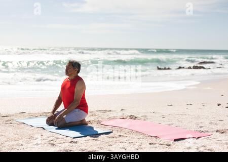 Senior man kneeling on blue yoga mat at sandy beach near shoreline rocks, copy space Stock Photo