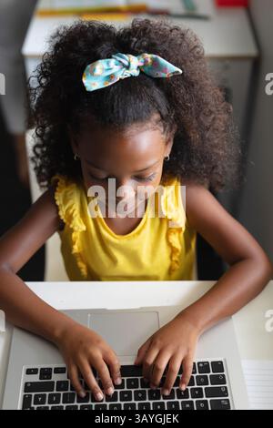 Child girl typing on silver laptop at home desk, showing focus with notebook and hair bow Stock Photo