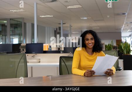 Mid-adult woman reviewing printed documents on conference table in open-plan office, copy space Stock Photo