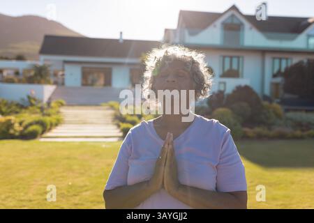 Senior woman standing on green lawn in front of suburban house, holding prayerful pose with glasses Stock Photo