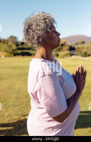 Senior woman meditating with hands in prayer gesture on grassy field, showing silver ring Stock Photo