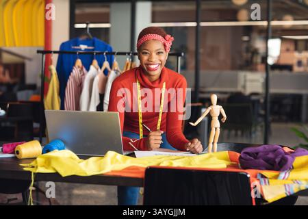African American woman sketching design on worktable in fashion studio, using measuring tape Stock Photo